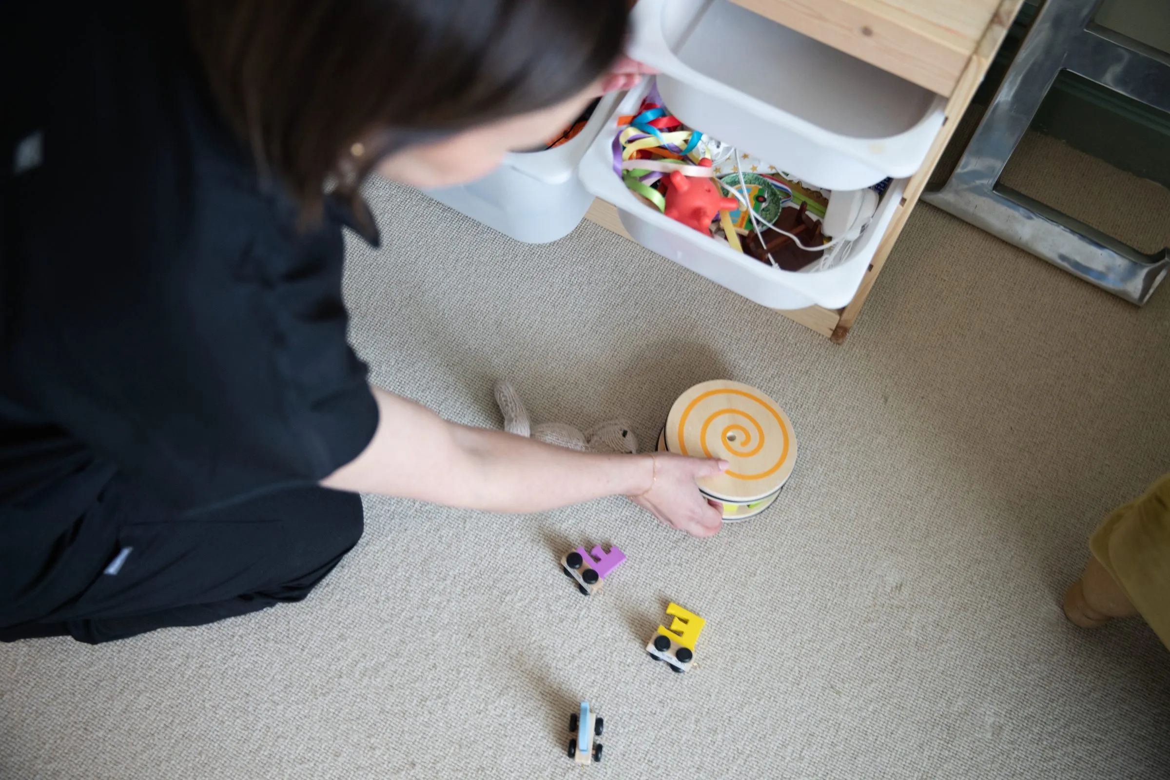 Overhead view of Grace kneeling on a carpet, gathering wooden toys into a basket.